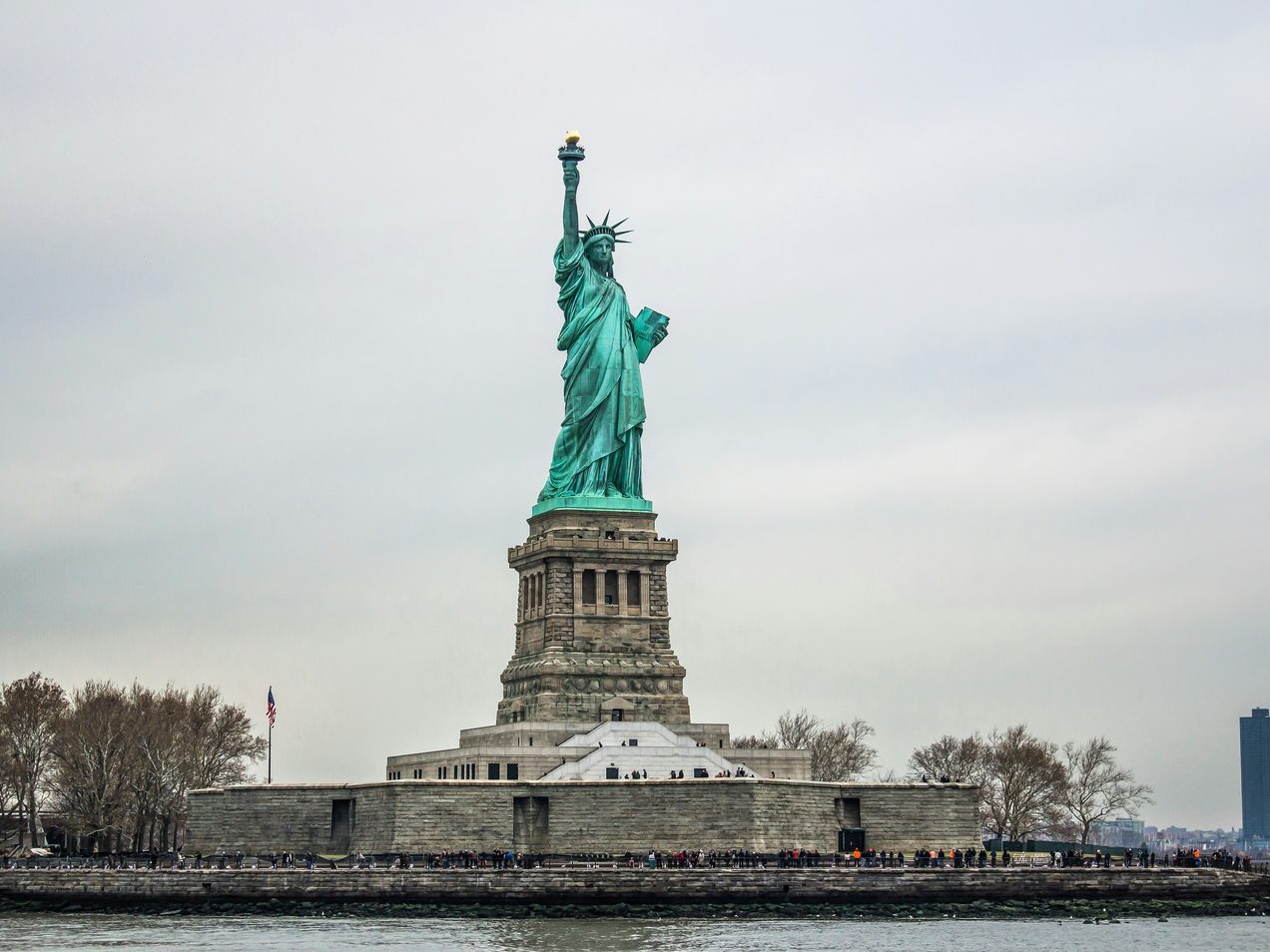 The Statue of Liberty seen from the water, visitors gathered along the sea wall below